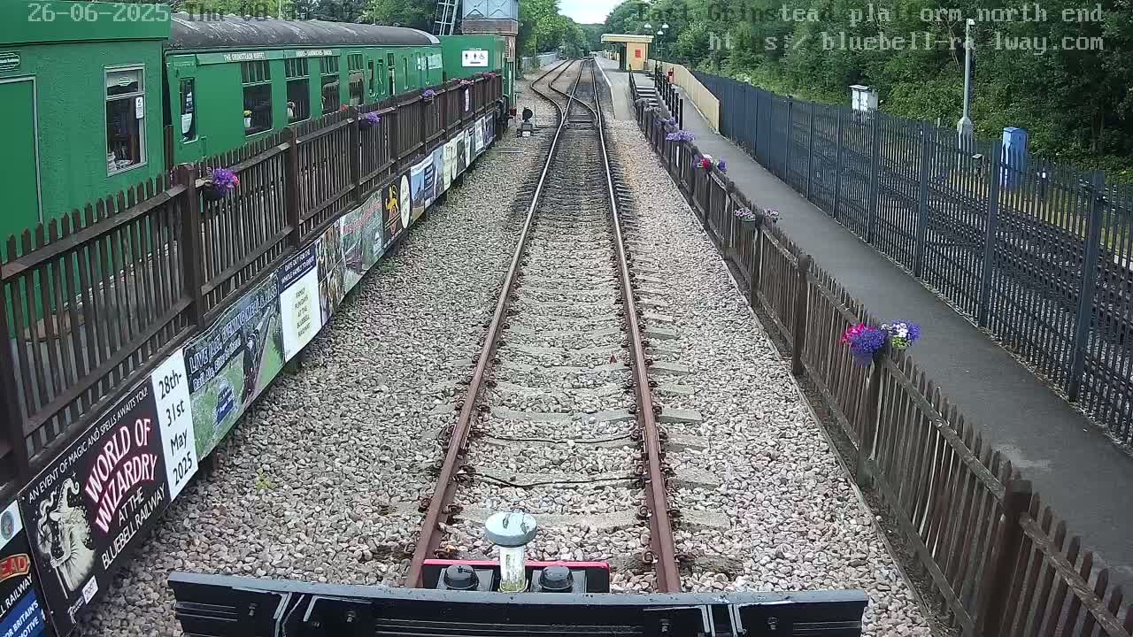 A view from a train's perspective shows railway tracks extending to a station platform lined with fences and flower pots, on a sunny day.