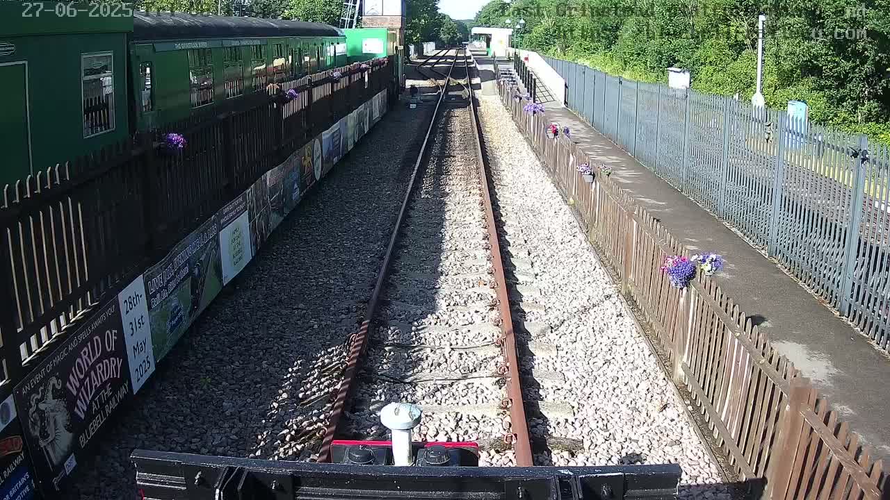 A sunny day at a railway station with a green train alongside a track lined with gravel and wooden fences decorated with flowers.