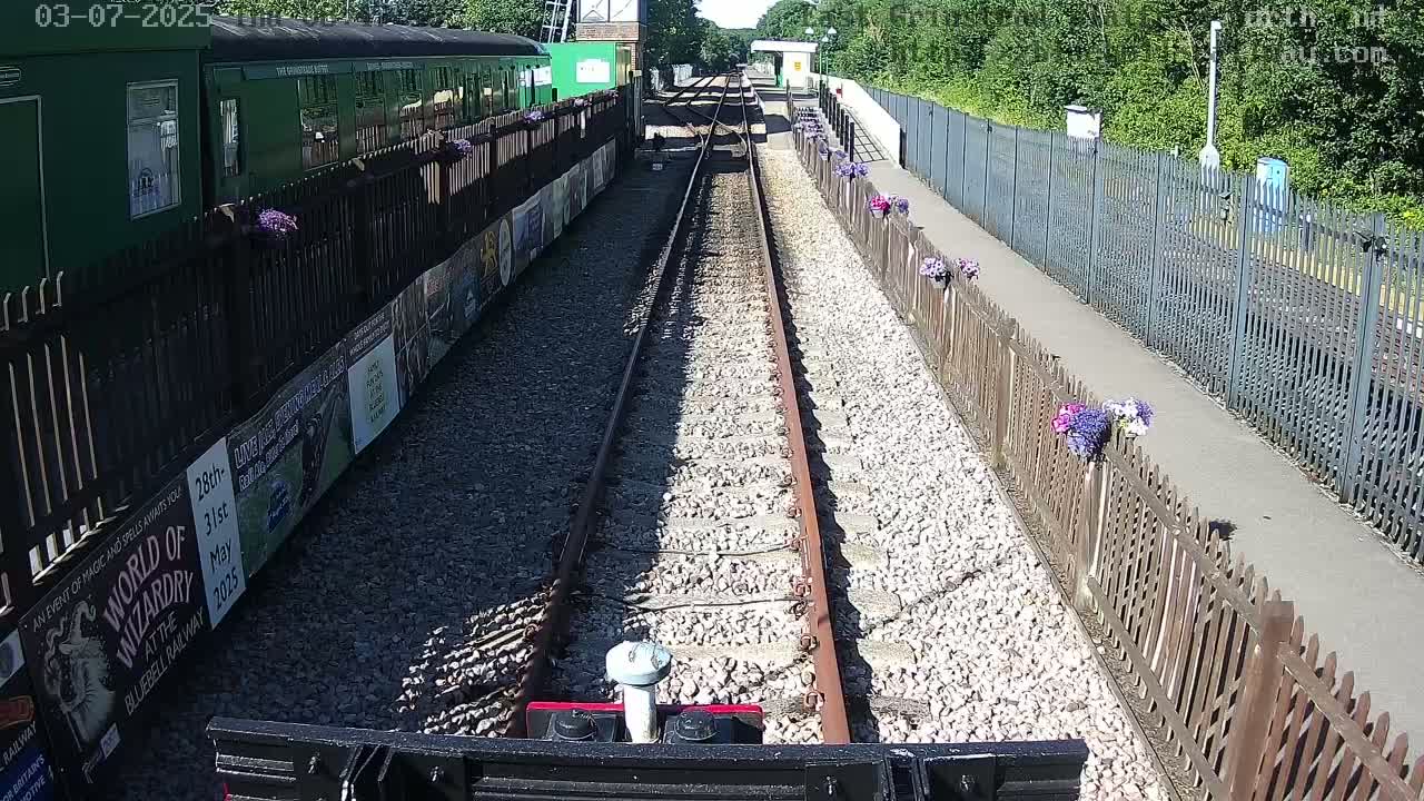 A train tracks lined with wooden fences and flowers stretches into the distance on a sunny day, with a green train visible to the side.