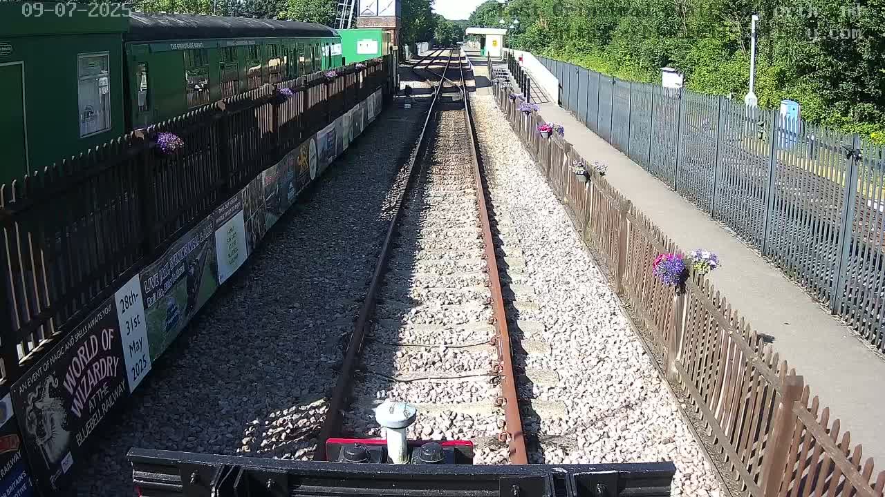 A train track lined with gravel and bordered by wooden fences with flower boxes stretches towards a green train car on a sunny day.
