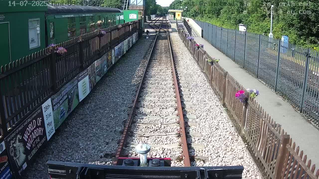 A view from a train's cab shows railway tracks leading to a station on a sunny day, with a green train and wooden fences lined with flowers alongside the tracks.