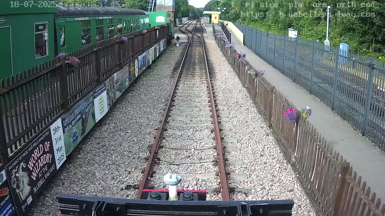 A train's point of view of a railway track leading to a station on a sunny day, with green train carriages and wooden fences lined with flowers along the platform.
