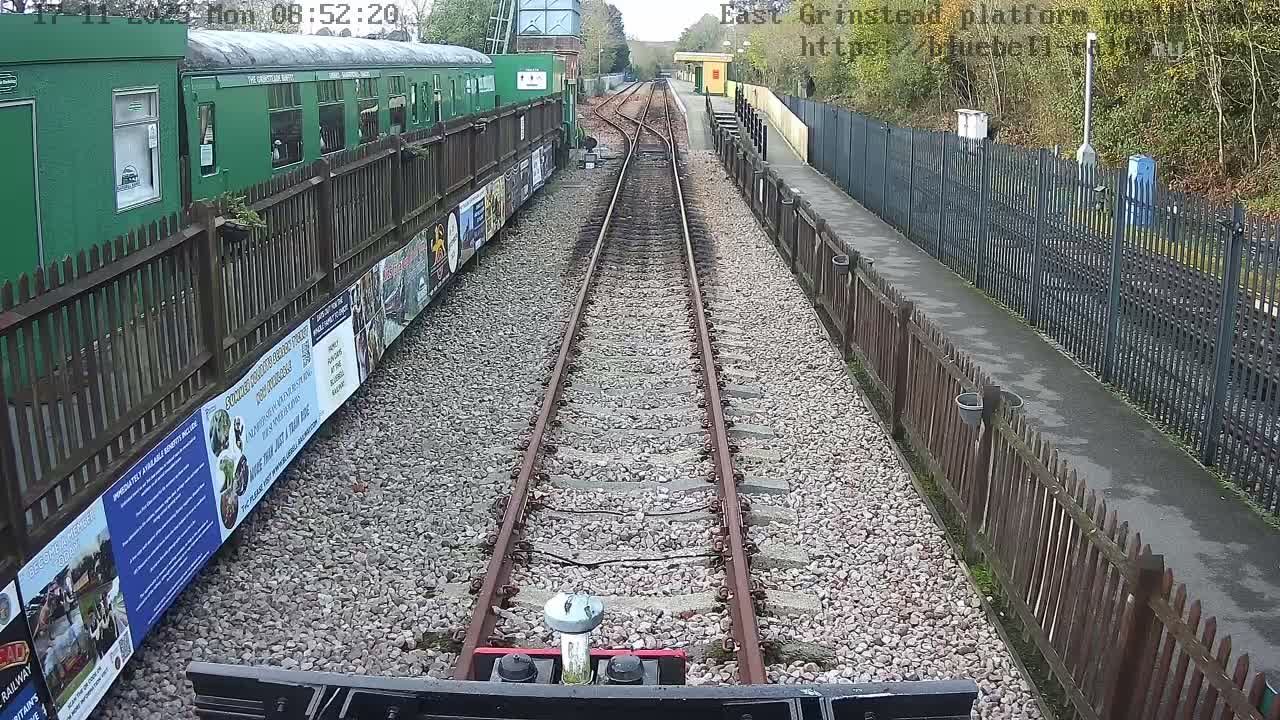 An outdoor train station is viewed from the tracks, showing two sets of rails converging in the distance, a long green train parked behind a wooden fence on the left, and a platform with a small yellow building behind another wooden fence on the right, all under an overcast sky.