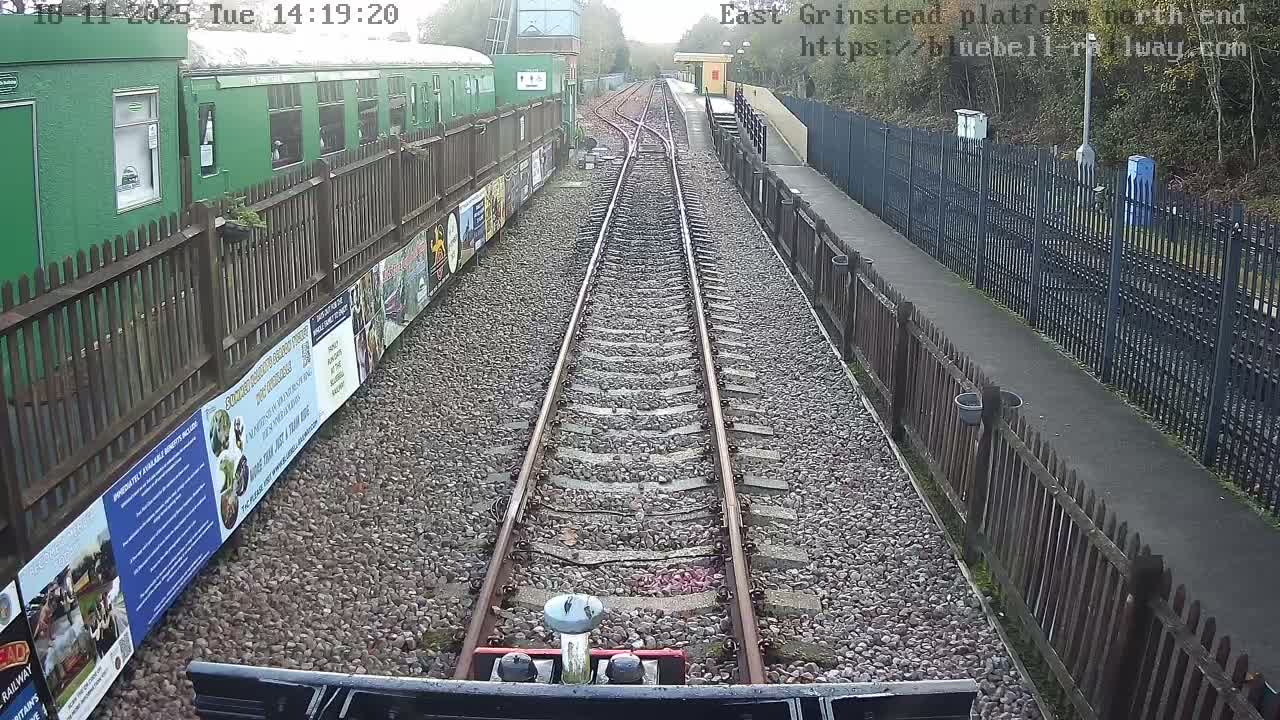 An outdoor train station is viewed from the tracks, showing two sets of rails converging in the distance, a long green train parked behind a wooden fence on the left, and a platform with a small yellow building behind another wooden fence on the right, all under an overcast sky.