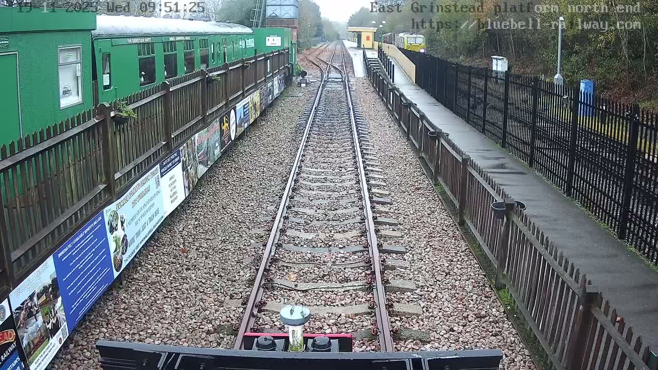 A pair of railway tracks recedes into the distance at a station, flanked by a green train on the left and a platform with a distant yellow building and partial yellow train on the right, all under overcast conditions.