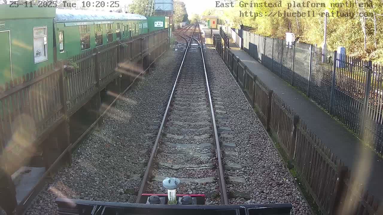 The image displays a sunny view down a double-track railway line from a station, with a green train carriage alongside a platform on the left and a fenced platform area on the right.