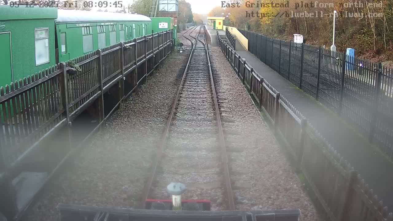 A view looking down two parallel train tracks at a railway station on an overcast day, with a green train car parked alongside a fenced platform on the left and another platform with a yellow building in the distance on the right.