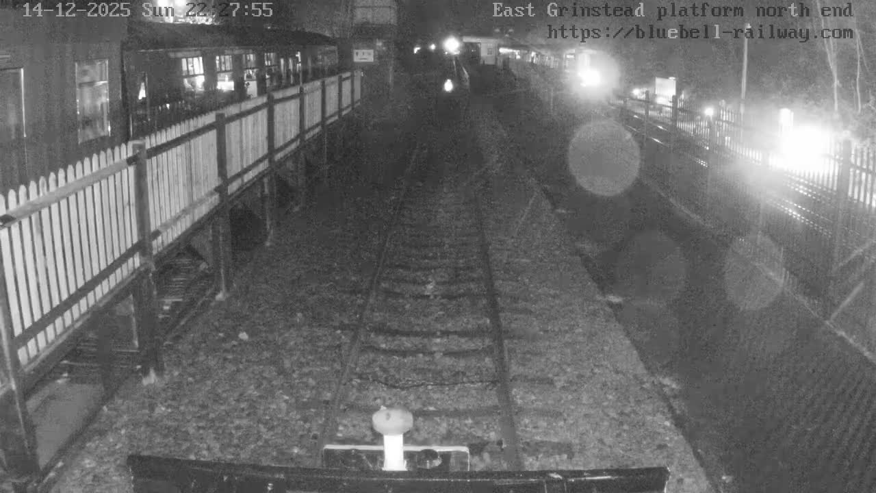 A view looking down two parallel train tracks at a railway station on an overcast day, with a green train car parked alongside a fenced platform on the left and another platform with a yellow building in the distance on the right.