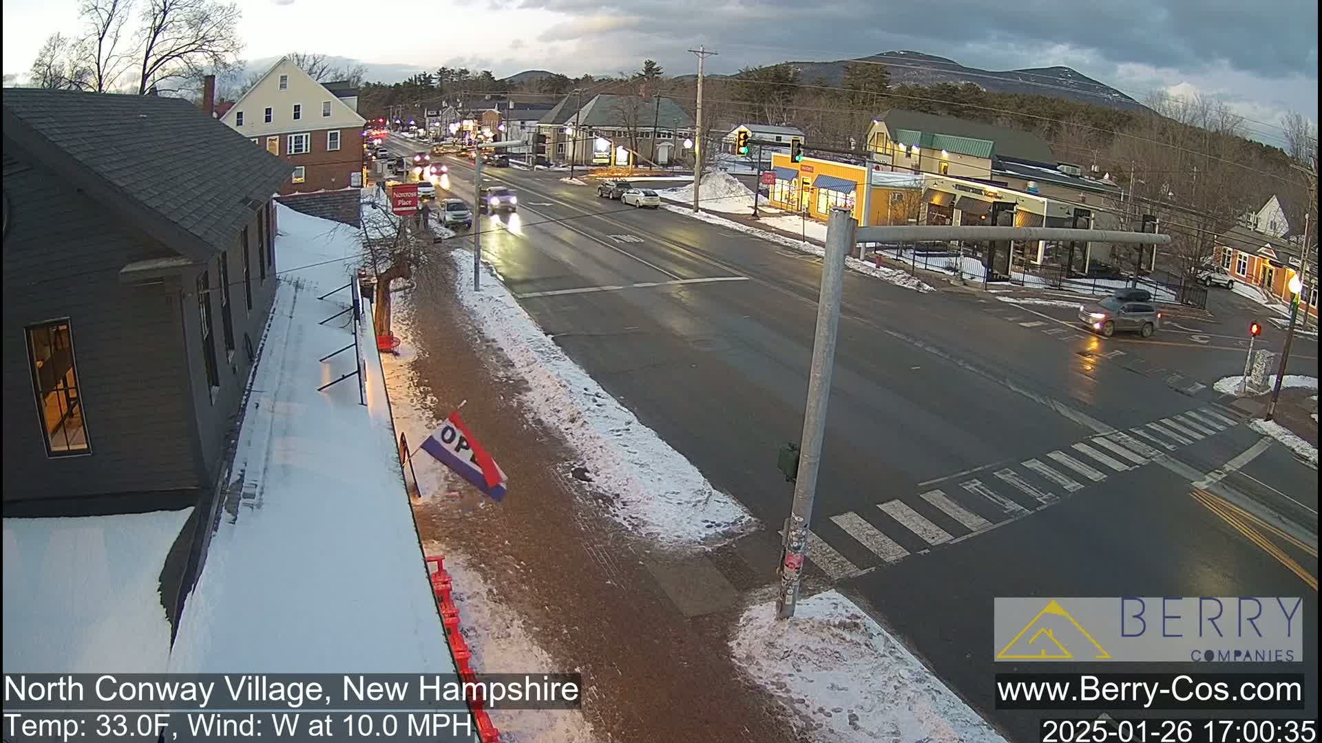 A street scene in a small town shows several cars driving on a wet road with patches of snow on the sides, under a partly cloudy sky.