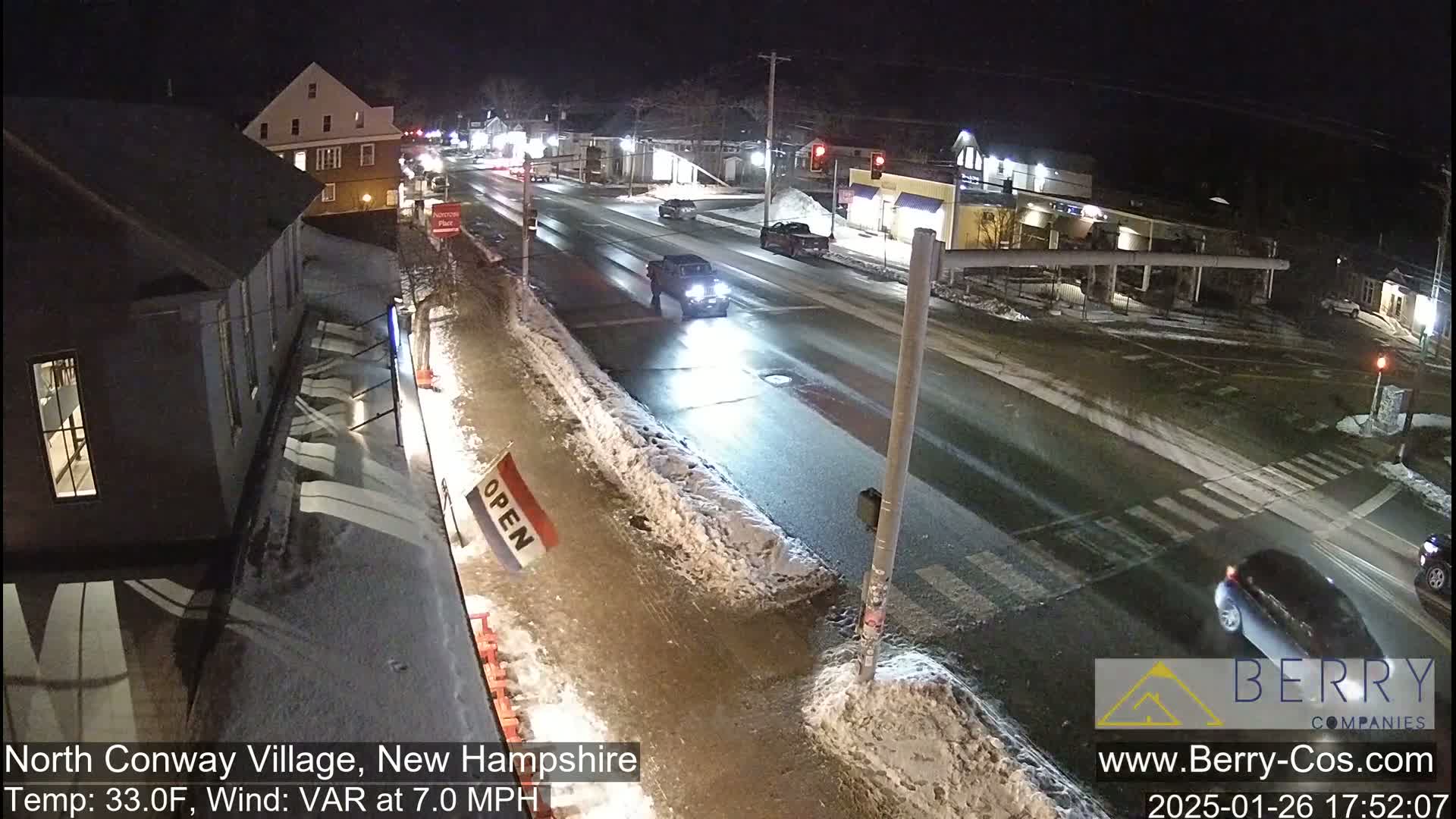 A nighttime street scene shows a sparsely trafficked intersection with snow on the sides of the road, under partly clear skies.