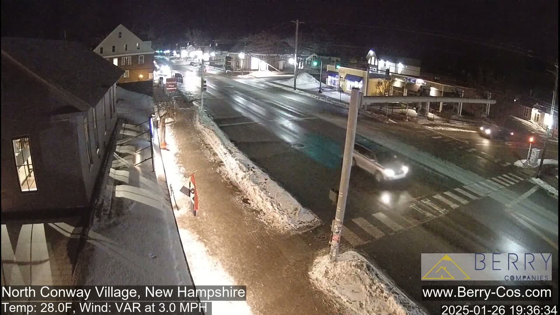 A nighttime street scene in a village shows a lightly trafficked road intersection with some snow and ice on the ground and a temperature of 28°F.