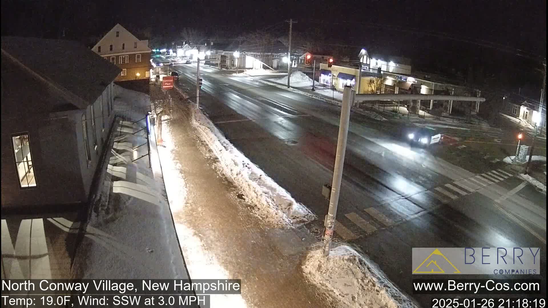 A nighttime street scene shows a mostly empty road with some snow on the ground and sidewalks, under a clear sky.