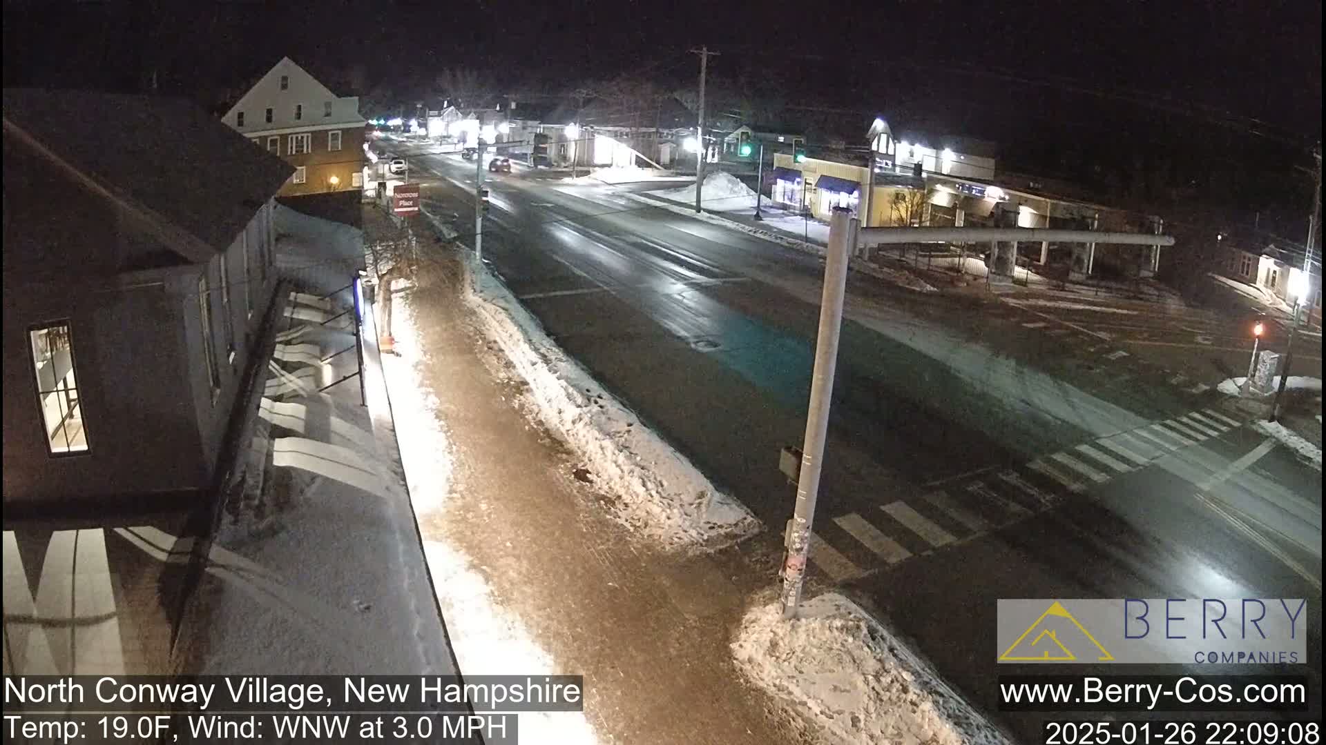 A snow-covered street intersection in a small town at night, with a few buildings and cars visible.