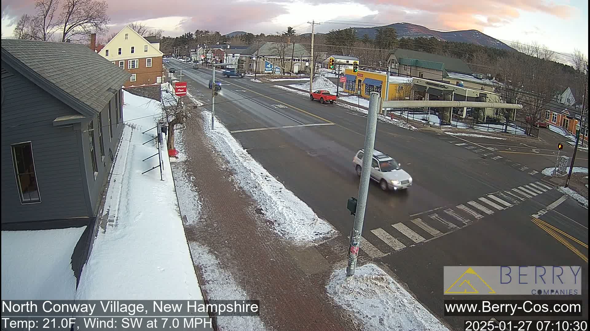 A snow-dusted street intersection in a small town shows several cars driving under a partly cloudy sunset sky.