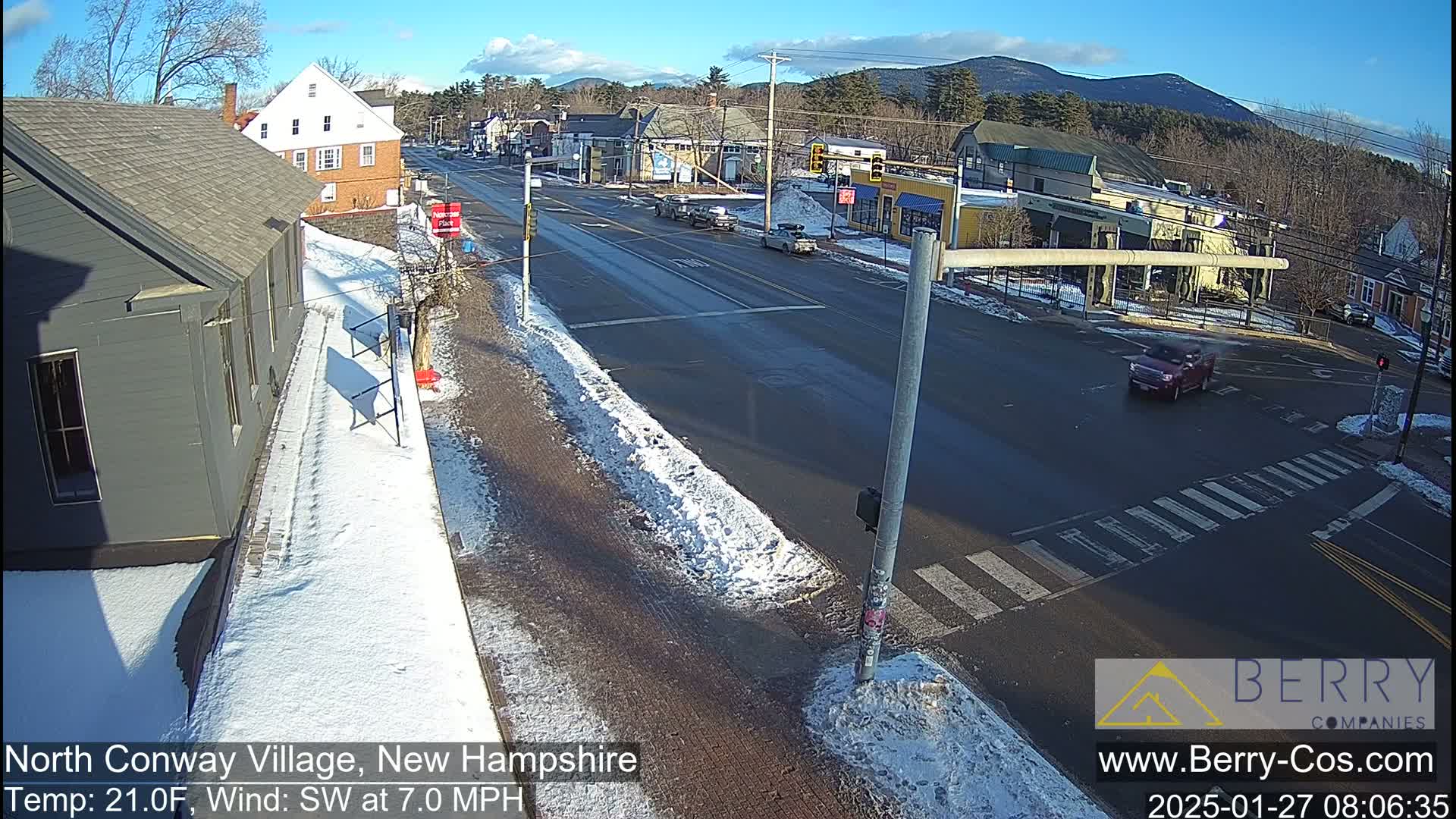 A partly sunny winter day shows a snow-covered street intersection with several cars and buildings in a village setting.