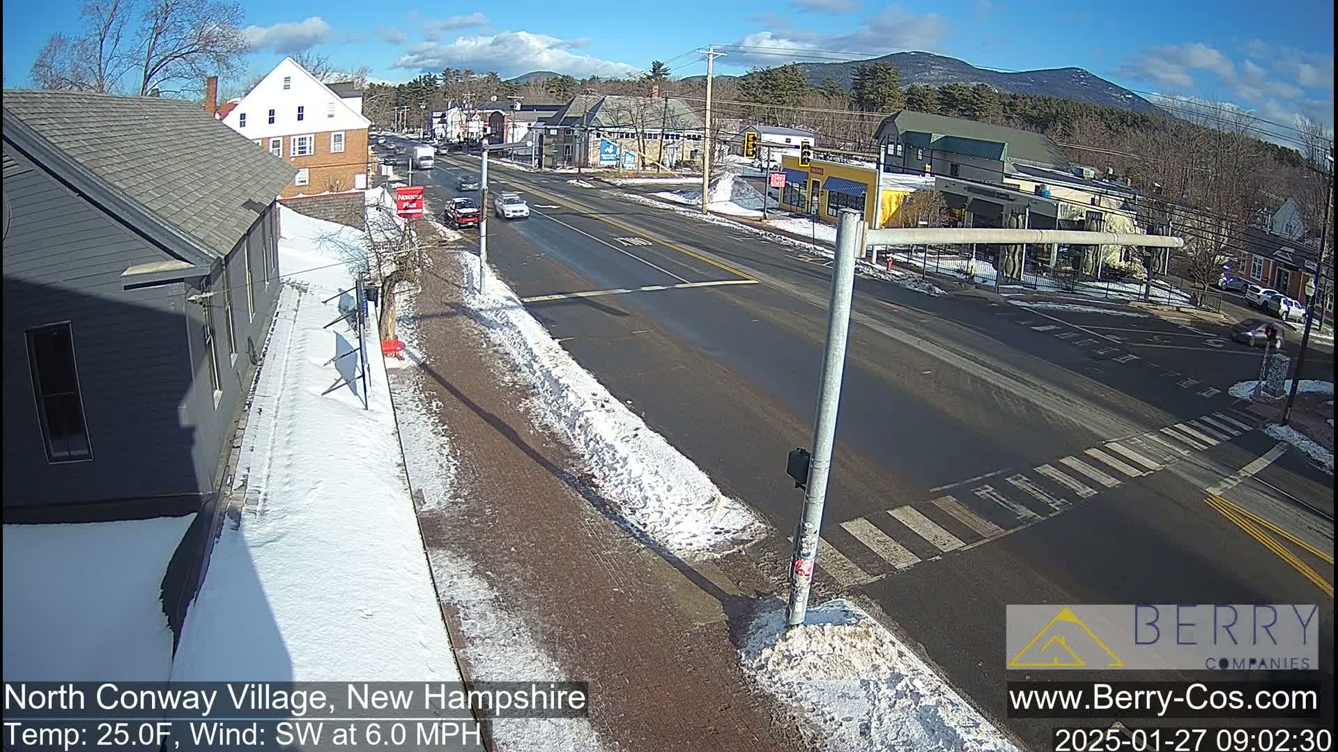 A sunny winter day shows a snow-covered street scene in a small town with several cars at an intersection,  mountains in the background.