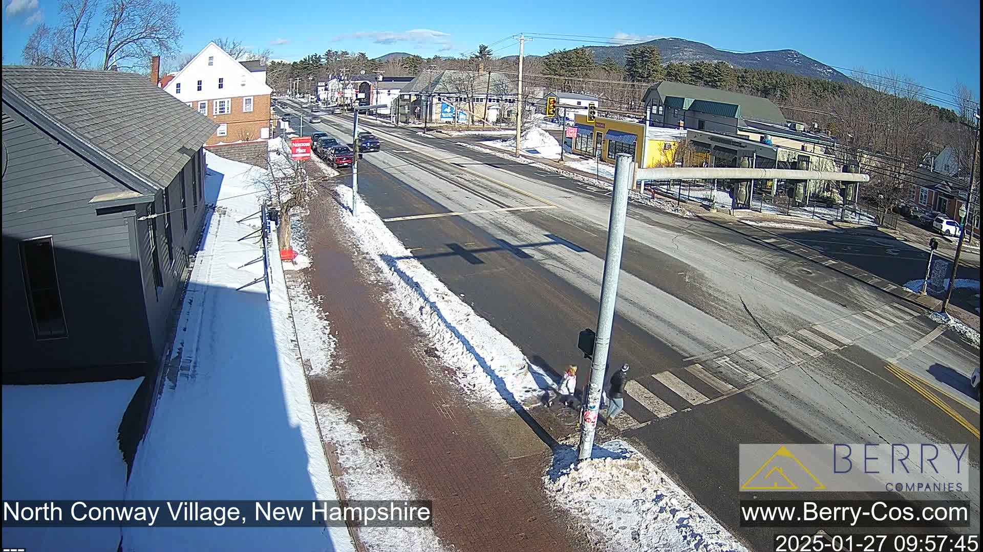 A sunny winter day in a town, featuring a snow-covered street intersection with pedestrians and a few cars, buildings line the street, and distant mountains are visible under a clear sky.