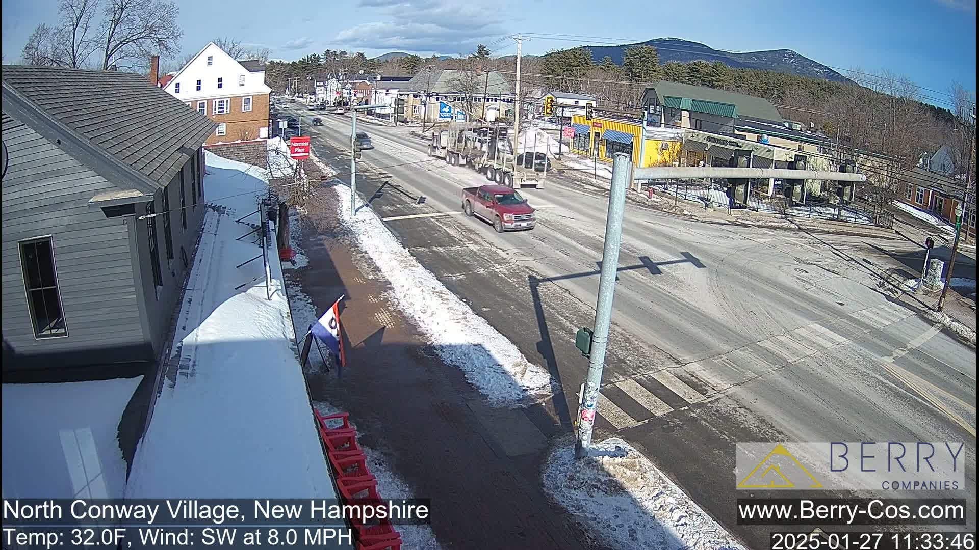 A sunny winter day shows a street scene with snow on the sidewalks and a few cars on the road, including a red pickup truck, with mountains in the background.