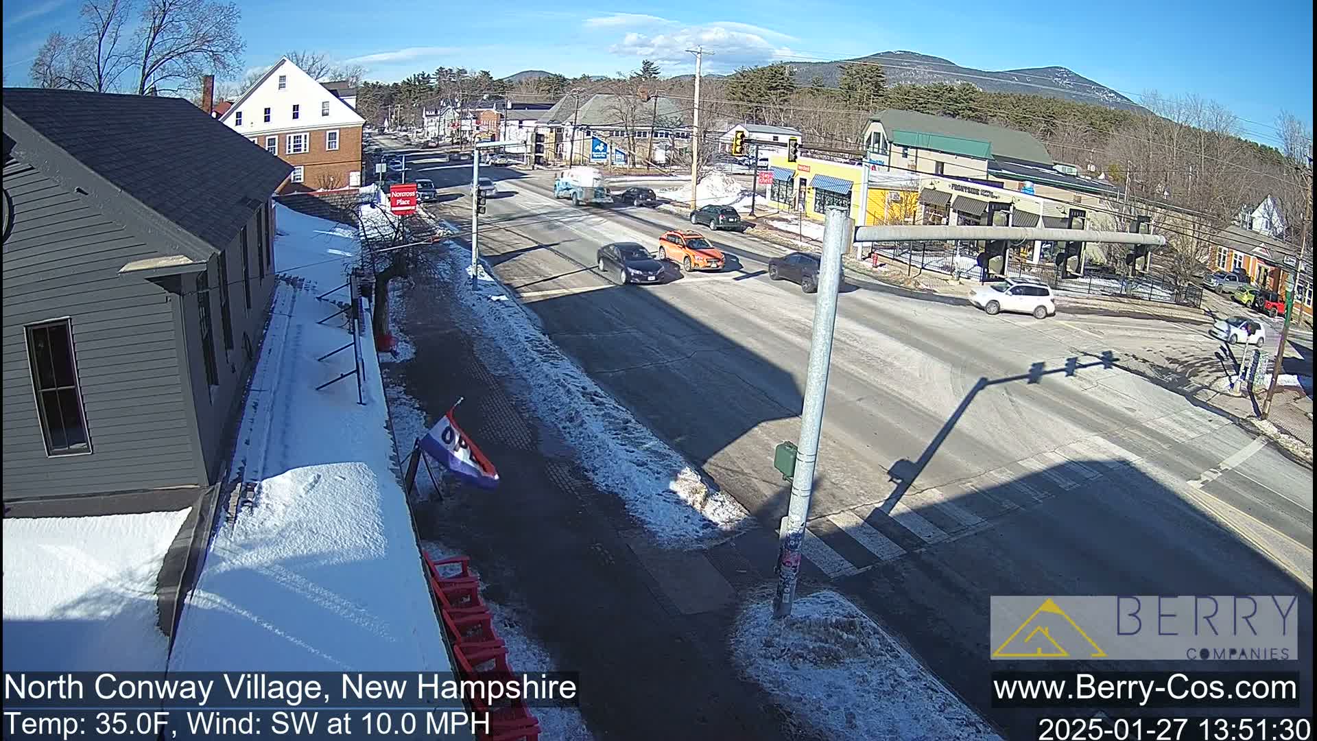 A sunny, snow-dusted street scene shows several cars at an intersection in a village, with mountains in the background and a light southwest wind.
