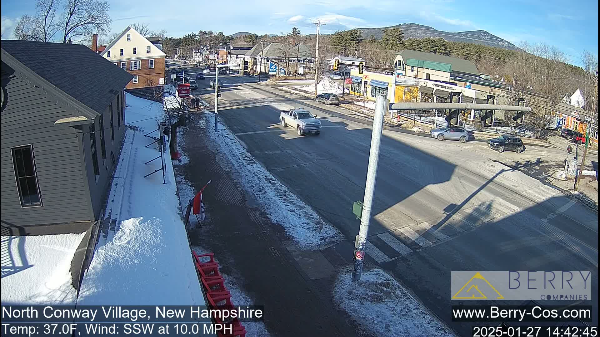 A sunny, snowy street scene shows a few cars at a four-way intersection in a village, with mountains in the background and snow on the ground.