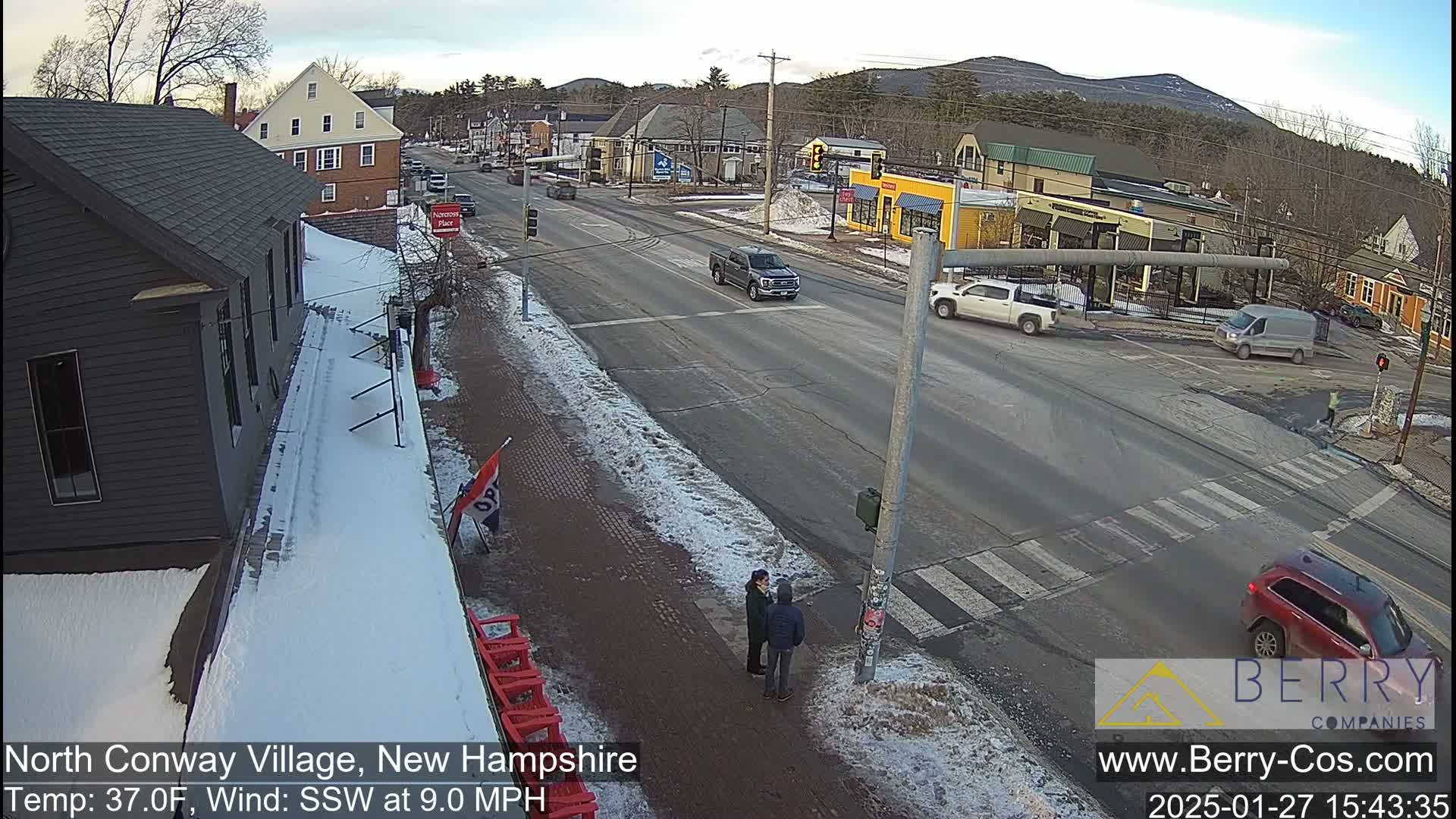 A snow-covered street scene shows a four-way intersection with several cars and pedestrians, under a partly cloudy sky.