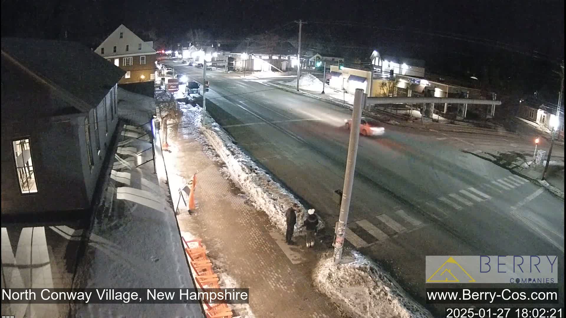 A nighttime street scene shows a snow-covered roadway with several cars and pedestrians, under dark, clear skies.
