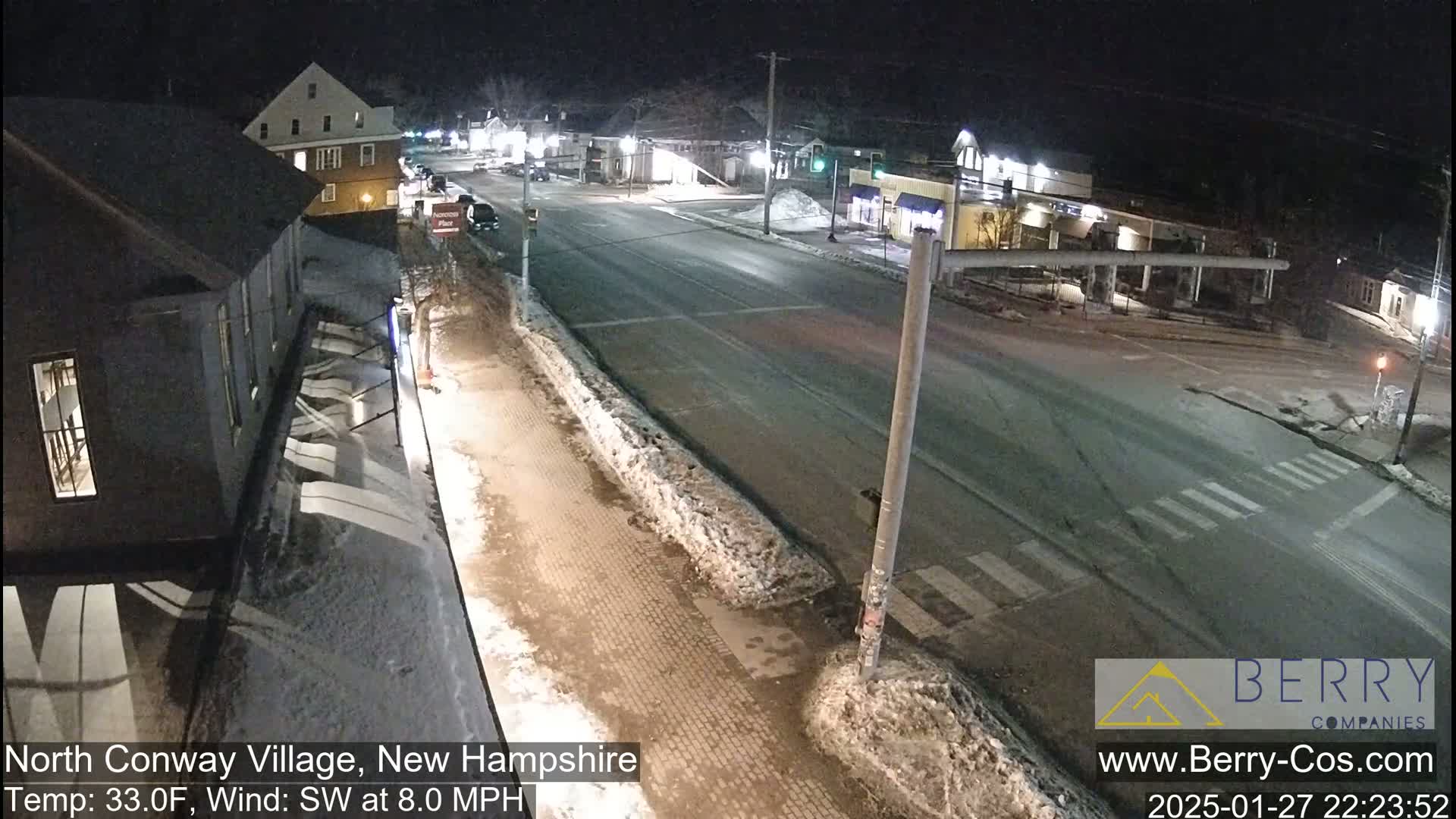 A nighttime street scene shows a snow-covered roadway and sidewalks with buildings lining the street under a clear sky.