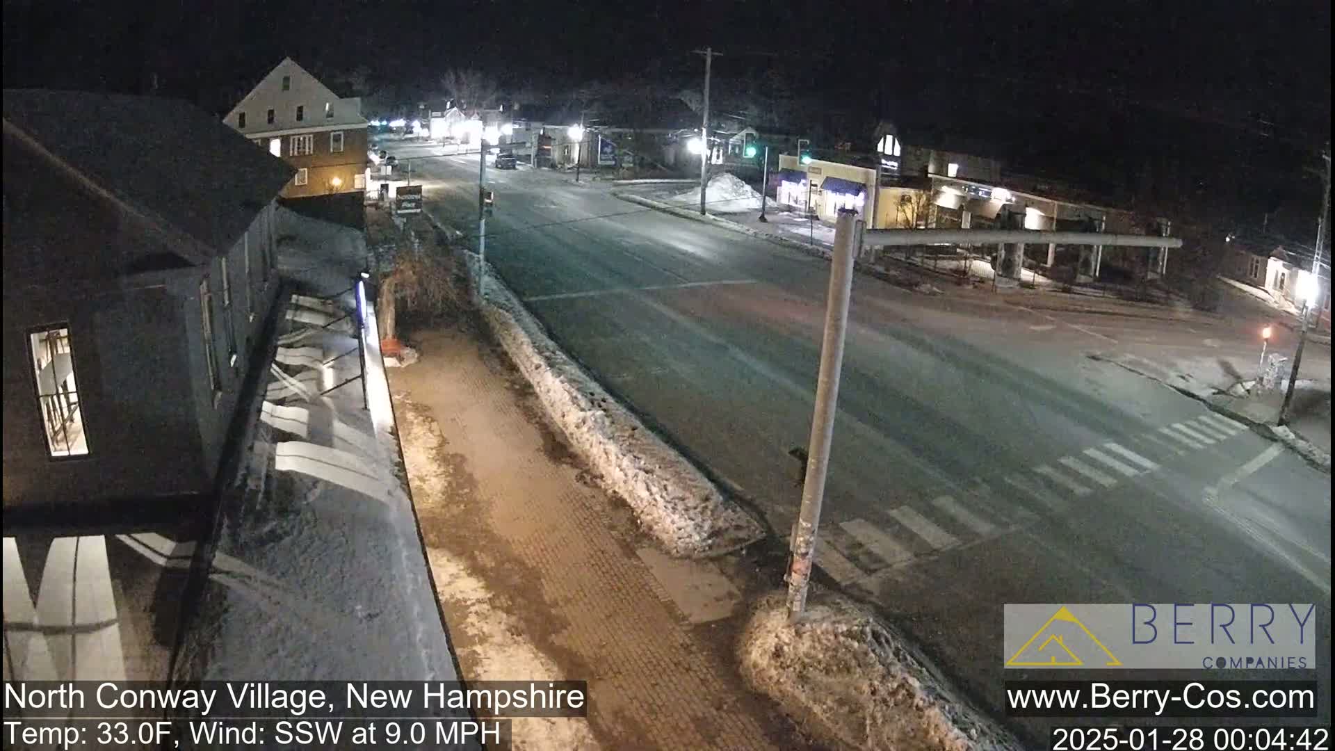 A nighttime street scene shows a mostly empty road intersection with some snow and ice on the ground, buildings lining the street, and a few cars.