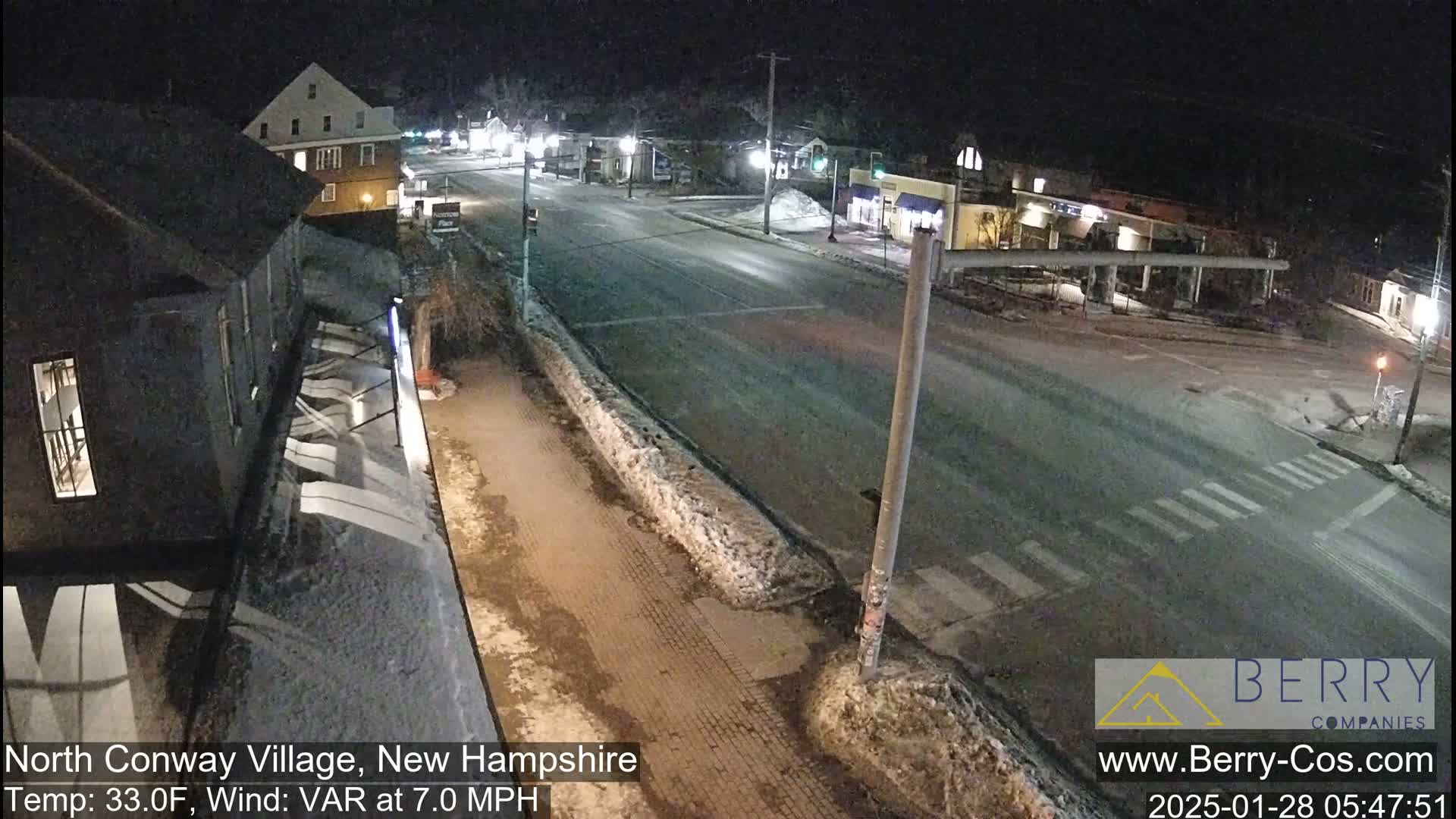 A nighttime street scene shows a mostly empty road with some snow and ice patches, a few buildings, and minimal traffic under partly cloudy conditions.