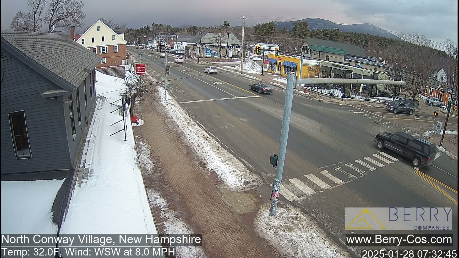 A snow-dusted street scene shows a town intersection with several vehicles, some snow on the sidewalks, and distant mountains under a cloudy sky.