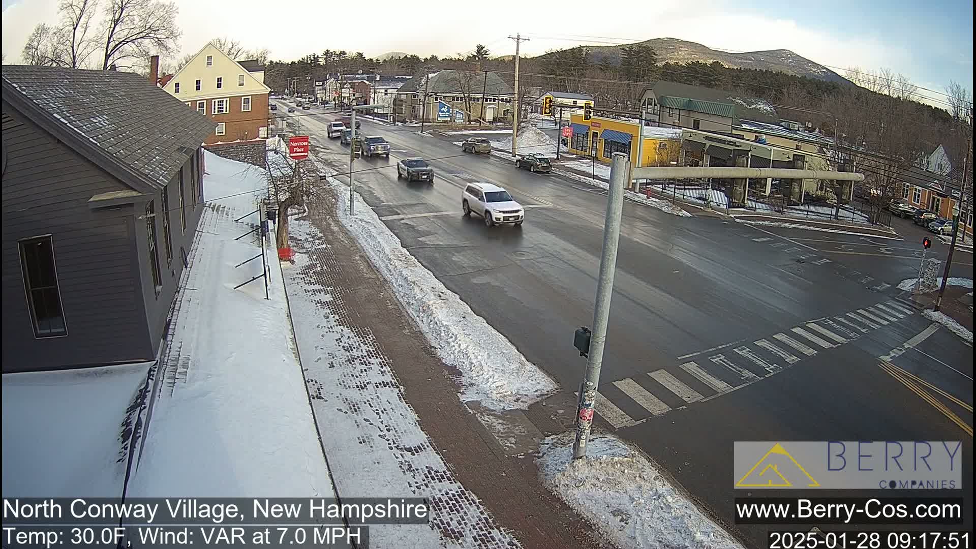 A partly snowy street scene shows several vehicles at an intersection, with a background of buildings and a mountain range under a mostly clear sky.