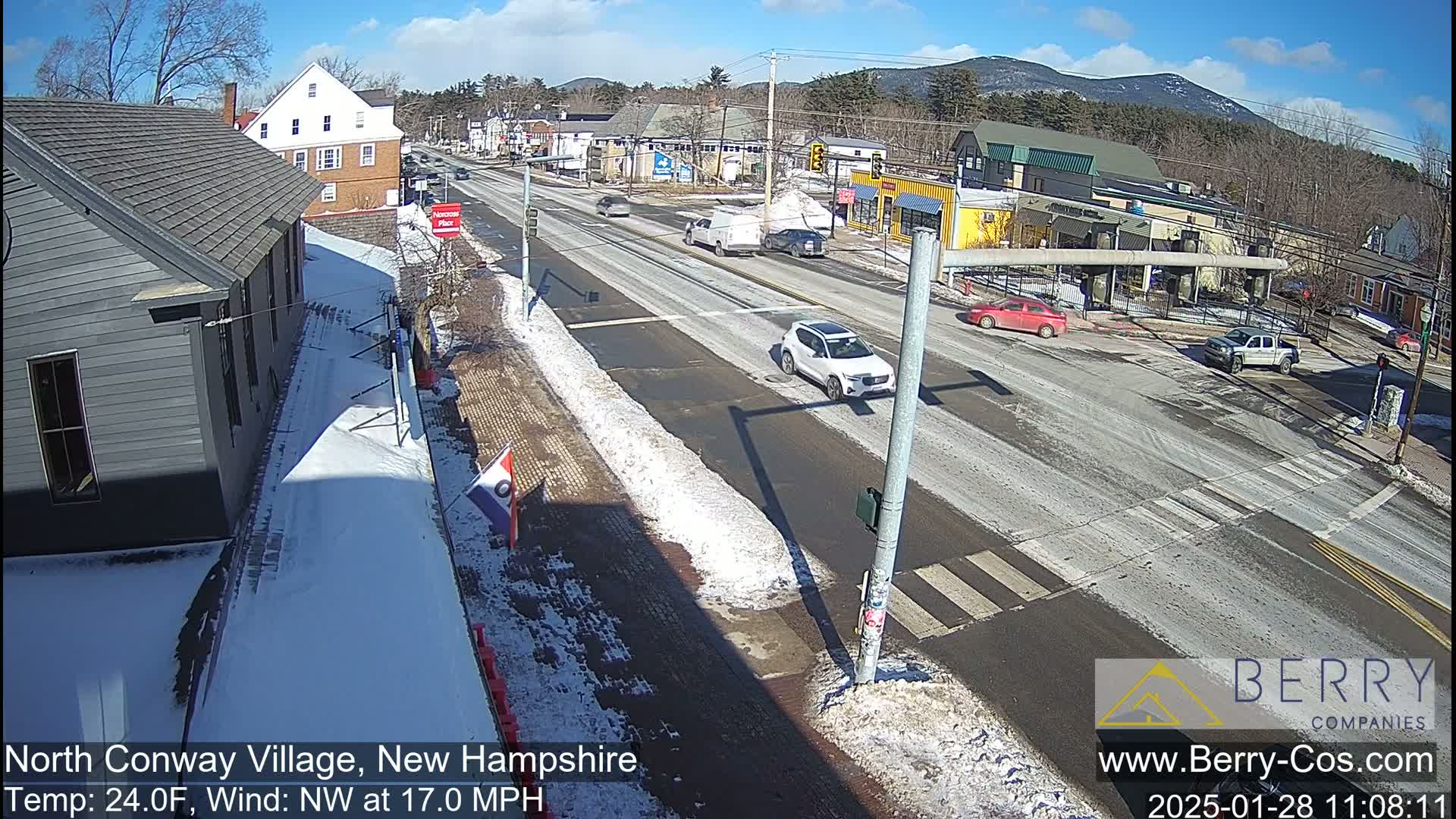 A sunny winter day reveals a snow-dusted street scene in a small town, with several cars at an intersection and buildings lining the road.