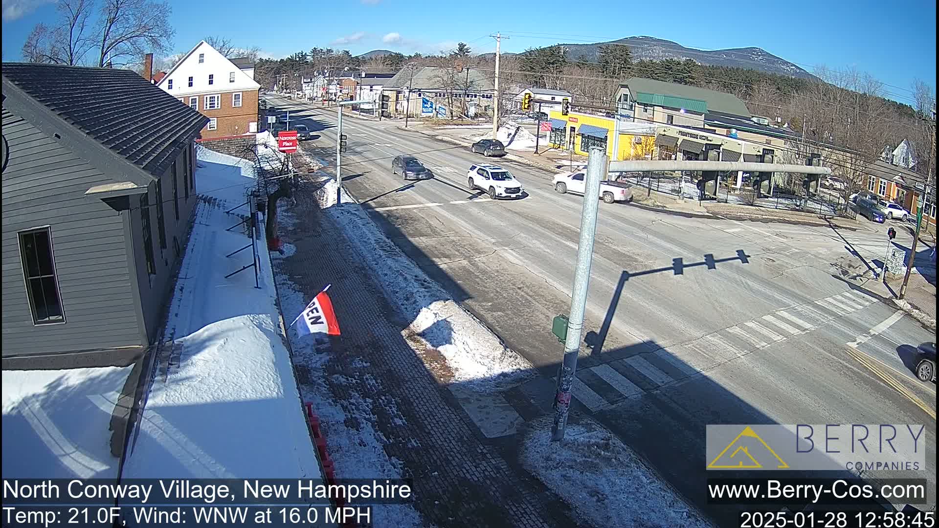 A sunny winter day shows a street scene with several cars at an intersection, snow on the sidewalks, and a few buildings visible in the background with mountains beyond.