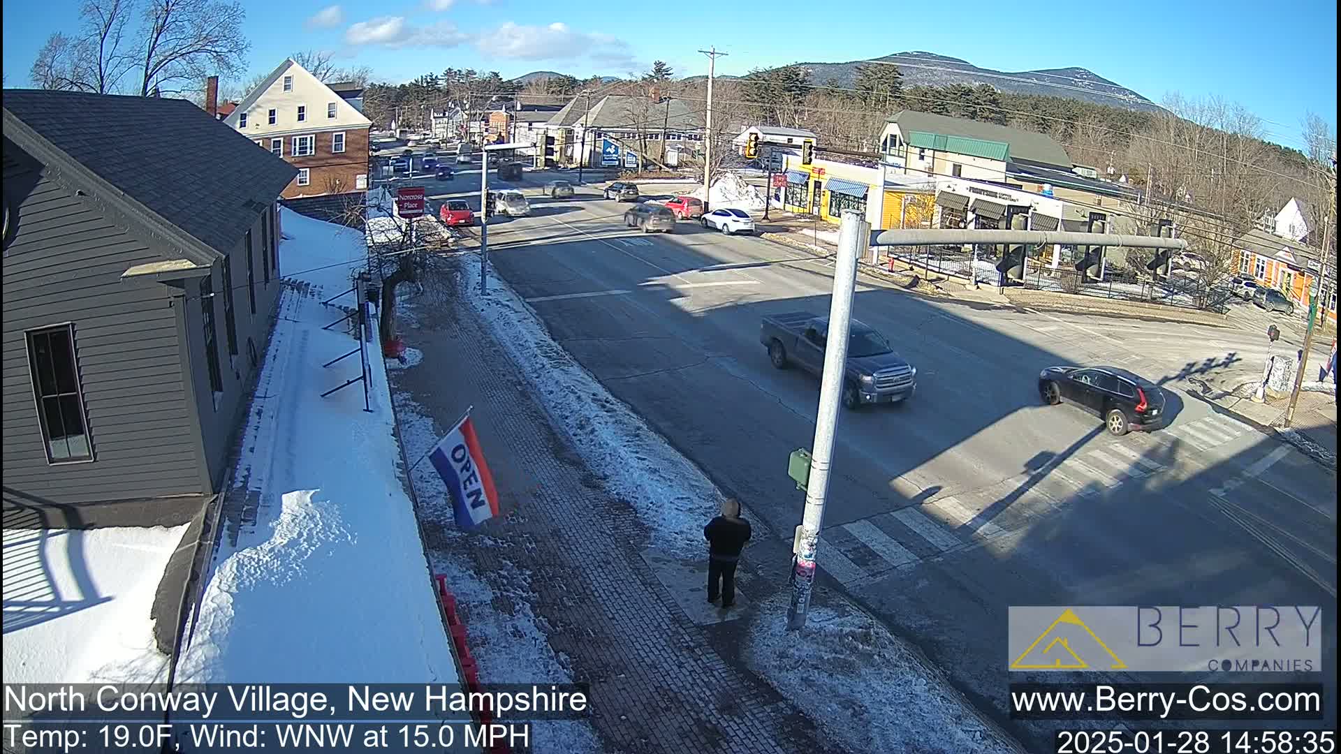 A sunny winter day shows a street intersection in a village with snow on the sidewalks and some cars driving by, with mountains in the background.