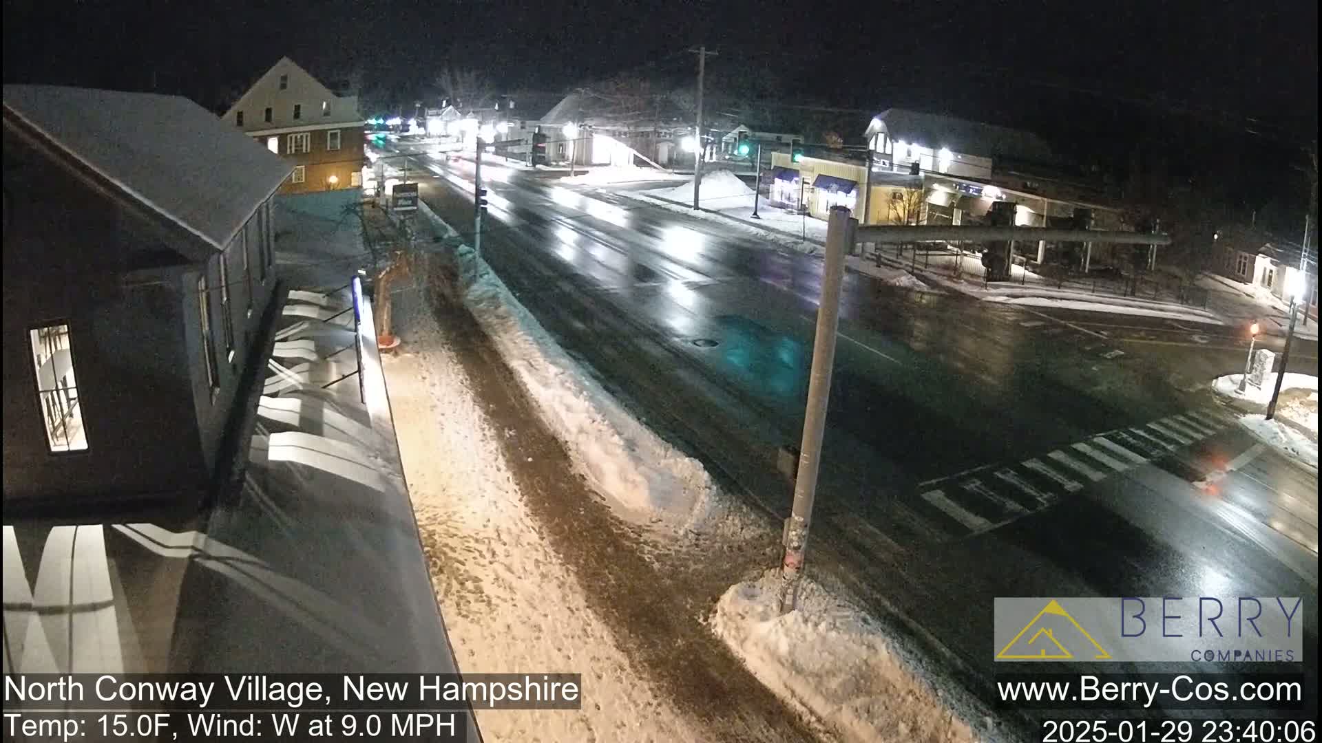 A wet, snow-covered street scene at night shows a mostly empty road with a few vehicles, flanked by buildings and some snow-covered sidewalks.
