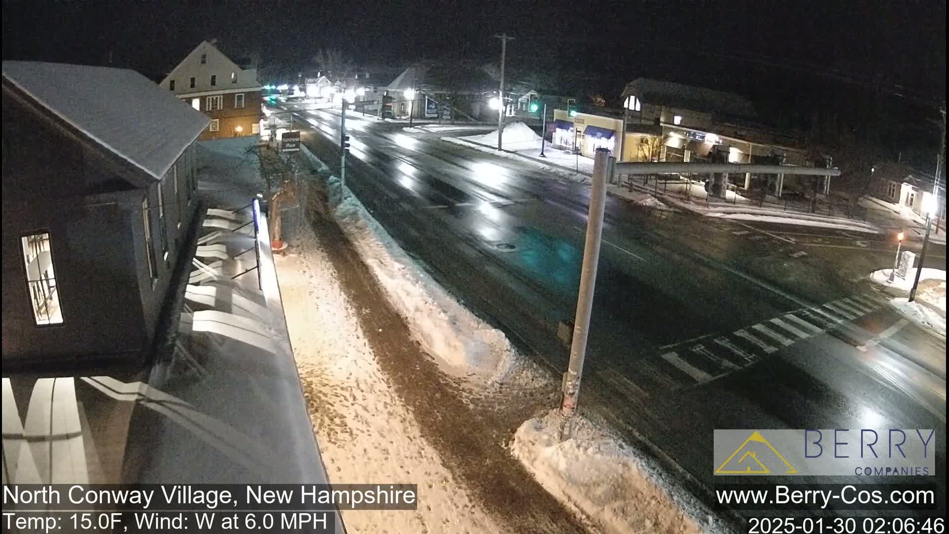 A nighttime view of a snow-covered street intersection in a village, with cars driving on a wet road.