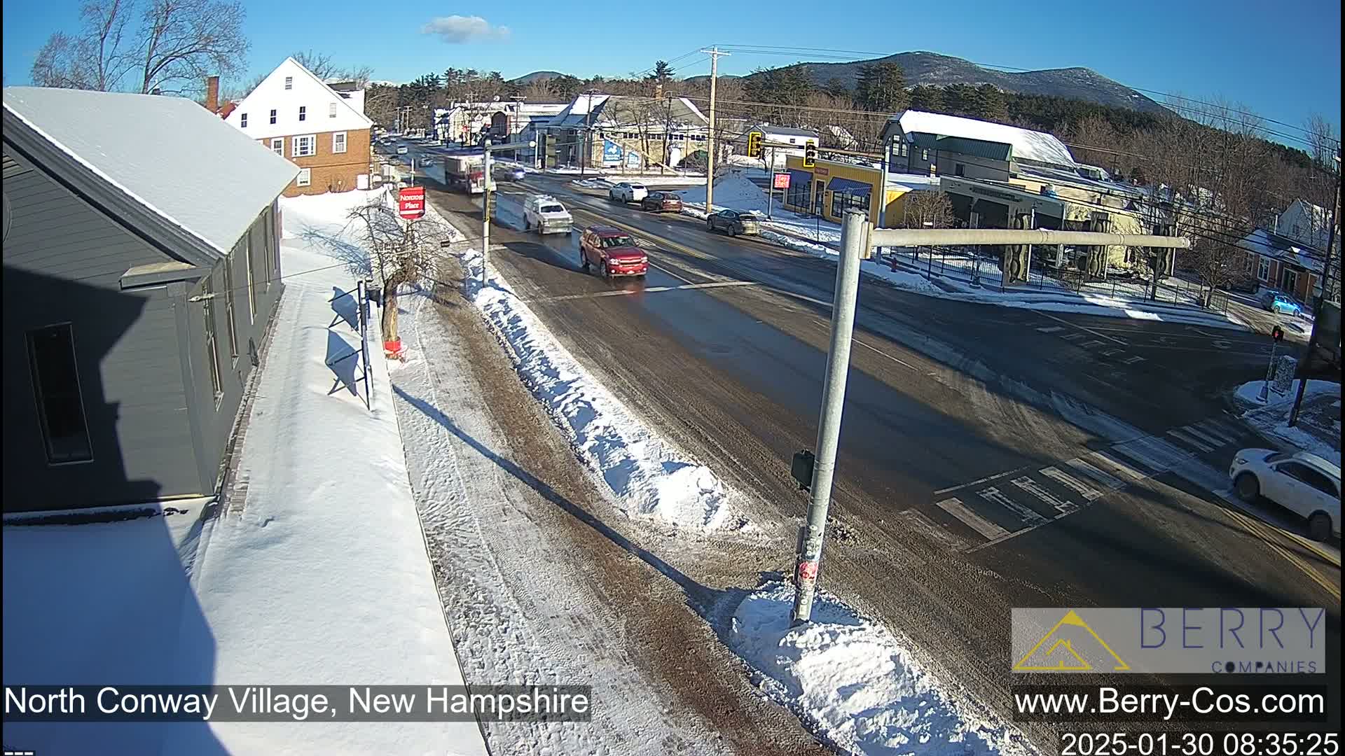 A sunny winter day shows a street scene with several cars driving on a road lined with snow banks, and buildings in the background.