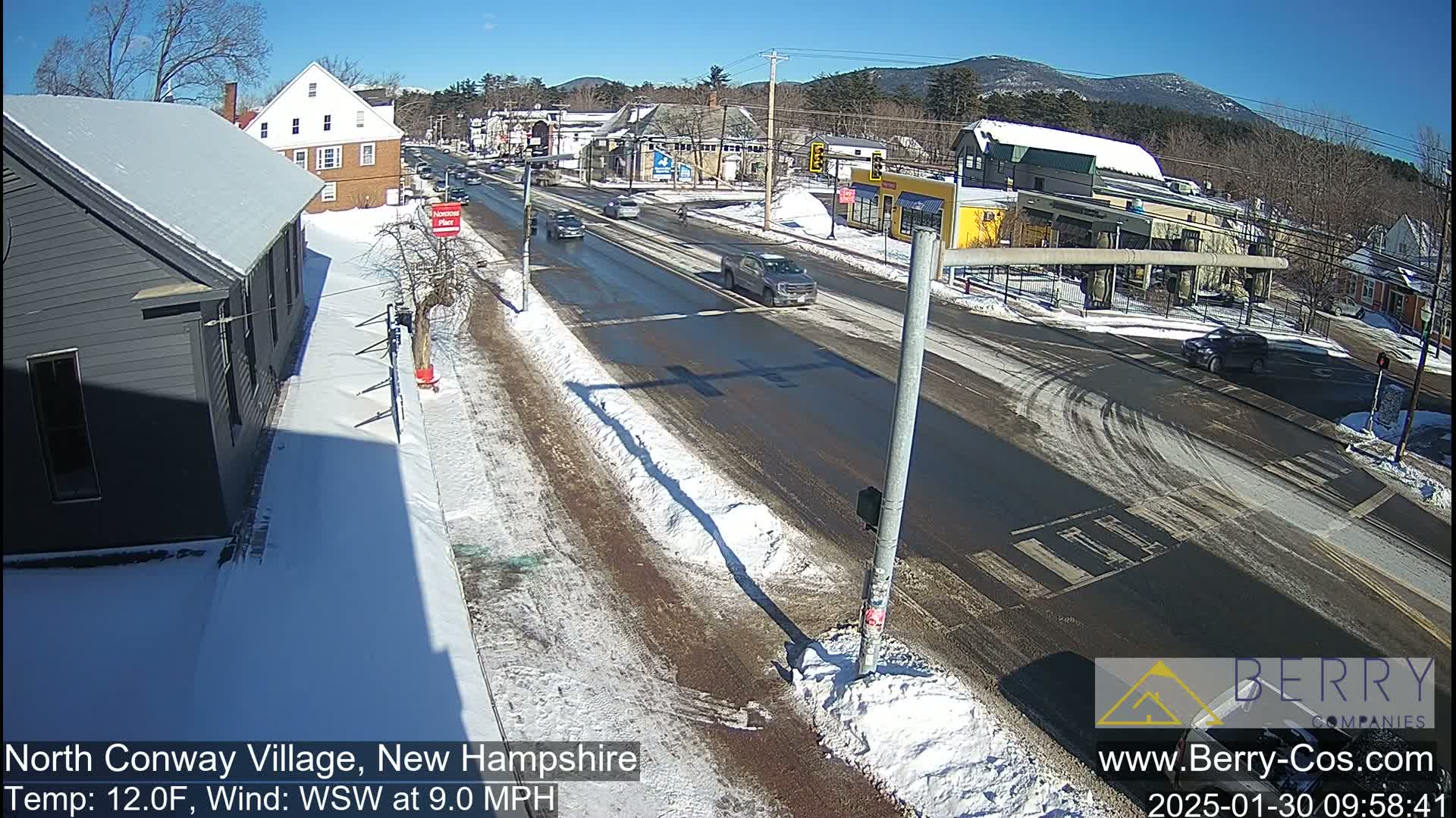 A sunny winter scene shows a snow-covered street intersection with several vehicles and buildings, under a clear blue sky.