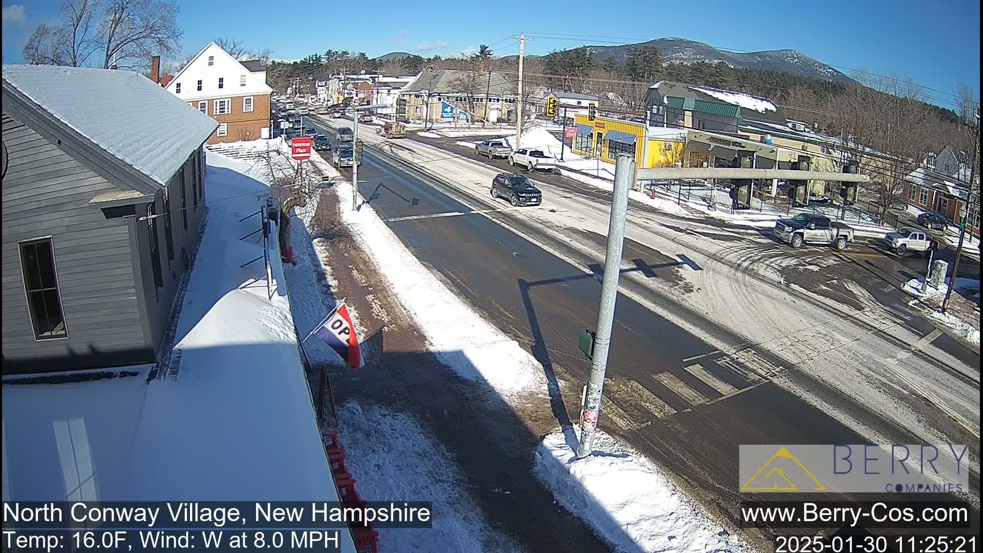 A sunny winter day scene shows a snow-covered street with cars and buildings, mountains in the background.