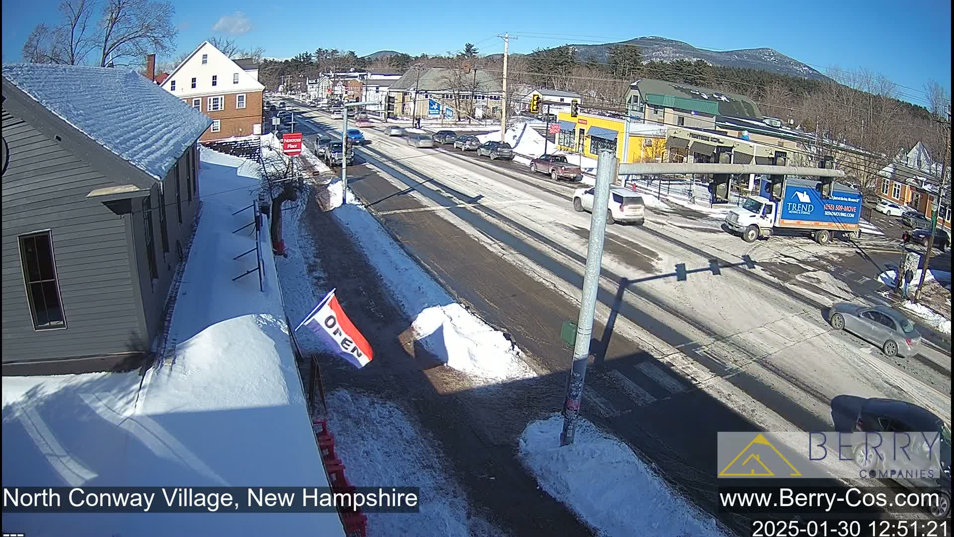 A sunny day in a village with snow on the ground and some cars driving on a street in front of buildings.