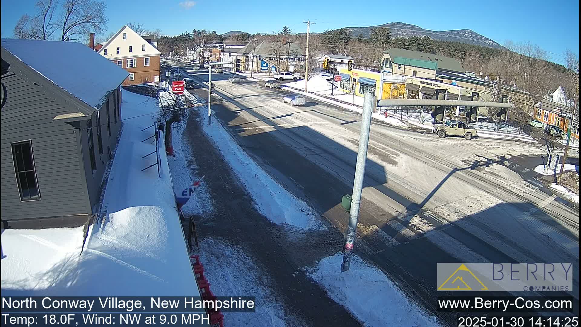 A snow-covered street scene shows a two-lane road with several cars, buildings, and some snow on the ground under a sunny sky.