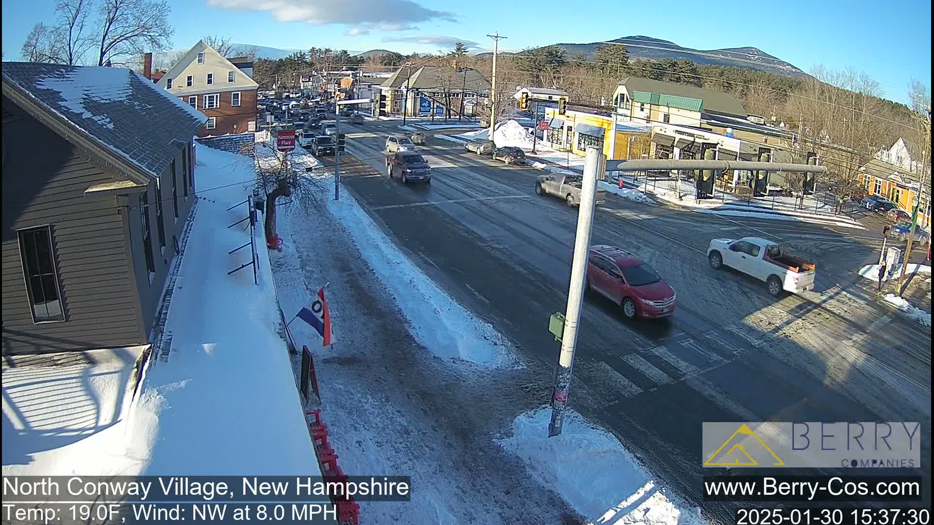 A snow-covered street scene shows a few cars at an intersection in front of some buildings, under a sunny sky.