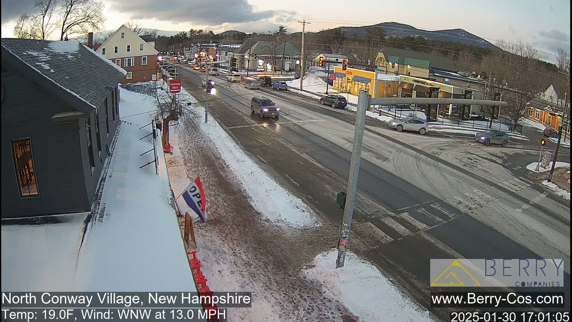 A snow-covered street scene shows a village with cars driving on a road that intersects with another road, under a partly cloudy sky.