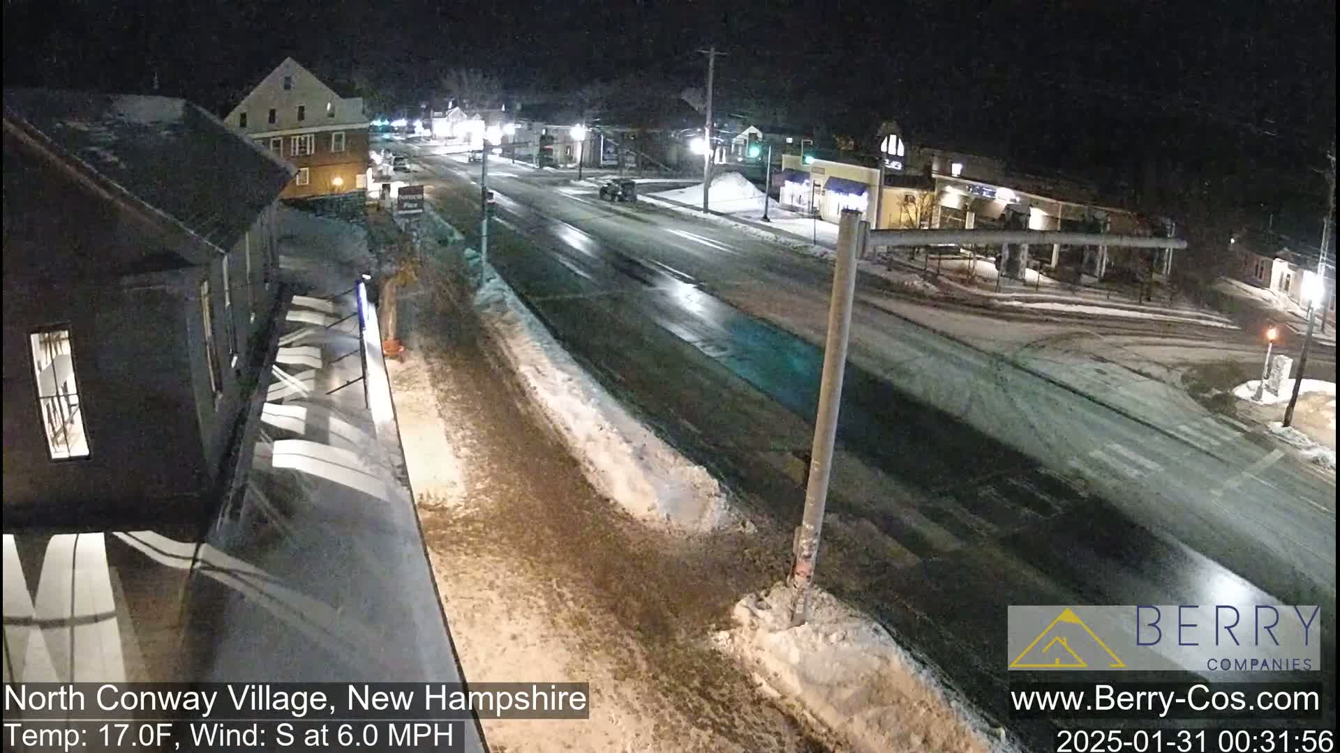 A nighttime street scene shows a mostly empty road with some snow and ice patches, a few cars, and buildings along the sides under partly cloudy conditions.