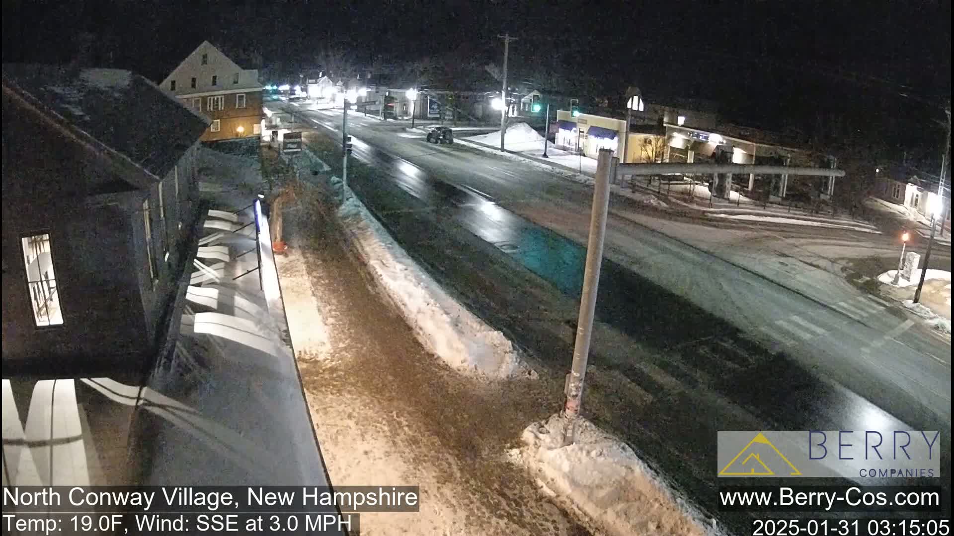 A nighttime view of a snow-dusted road in a village, with some buildings and cars visible under a dark sky.