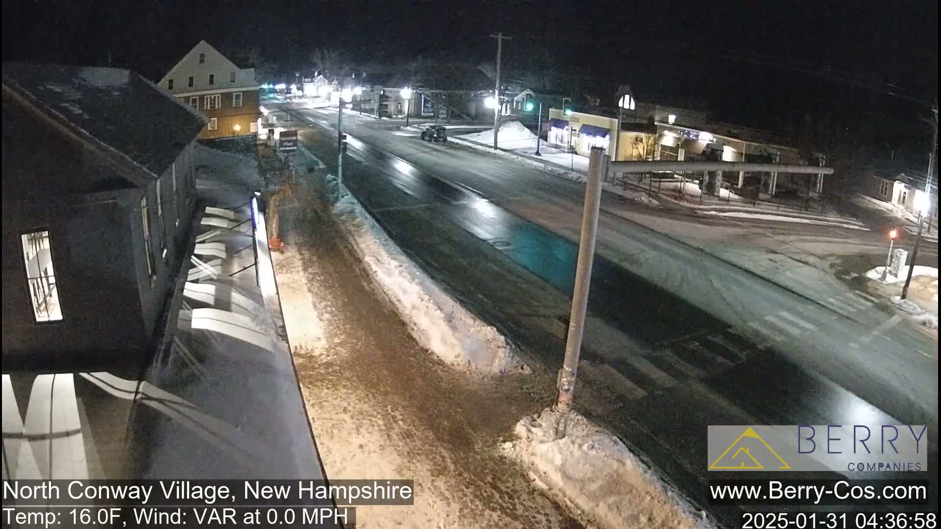 A nighttime street scene shows a mostly empty road with some snow and ice patches on the sides, a few cars, and buildings along the road.