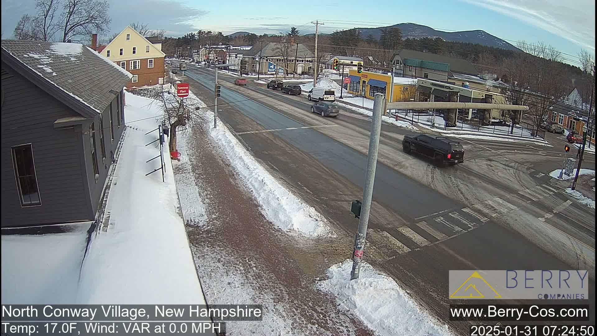 A snow-covered street scene in a small town shows several vehicles at a multi-way intersection on a partly cloudy day.