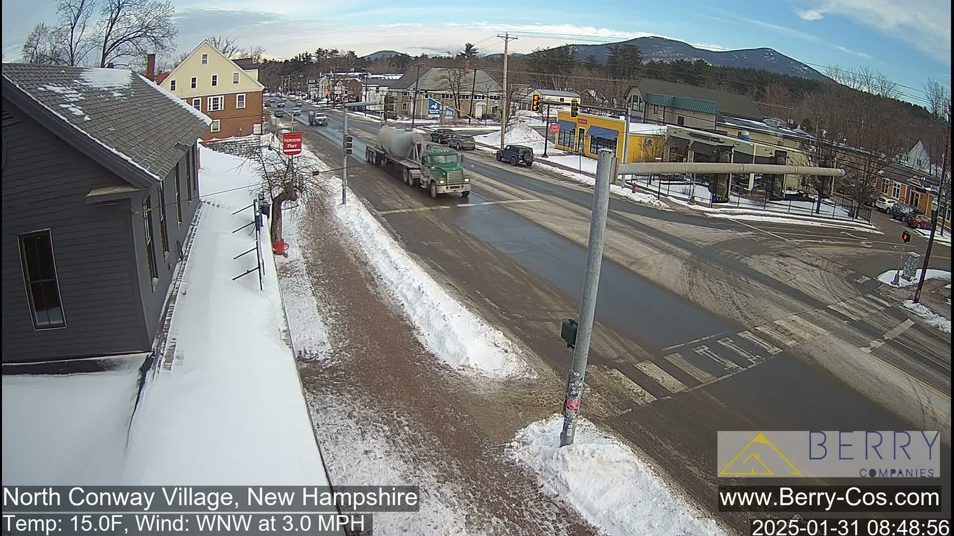 A semi-truck drives down a snow-covered road in a village, with several other vehicles and buildings visible under a clear sky.