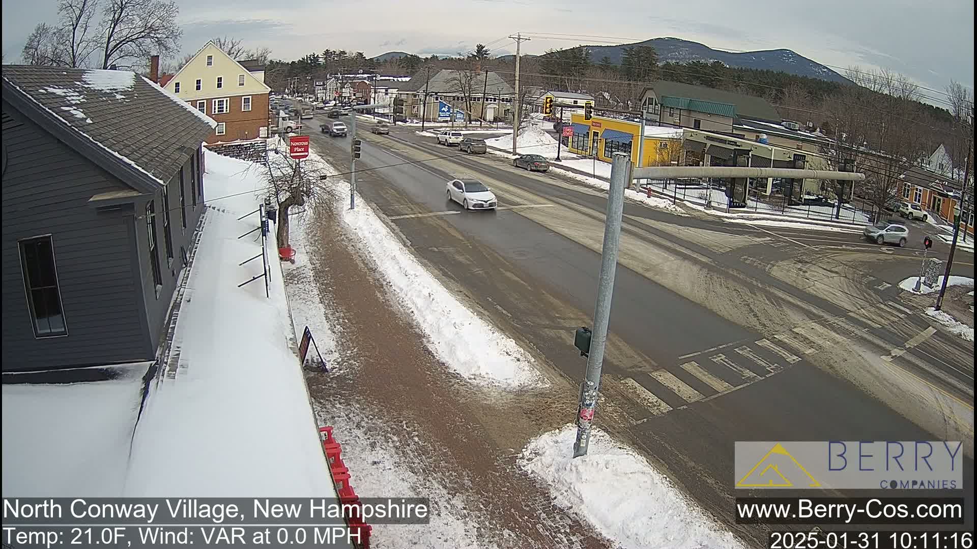 A snow-covered street scene shows a four-way intersection with several cars, buildings, and some snow on the ground in a partly cloudy day.