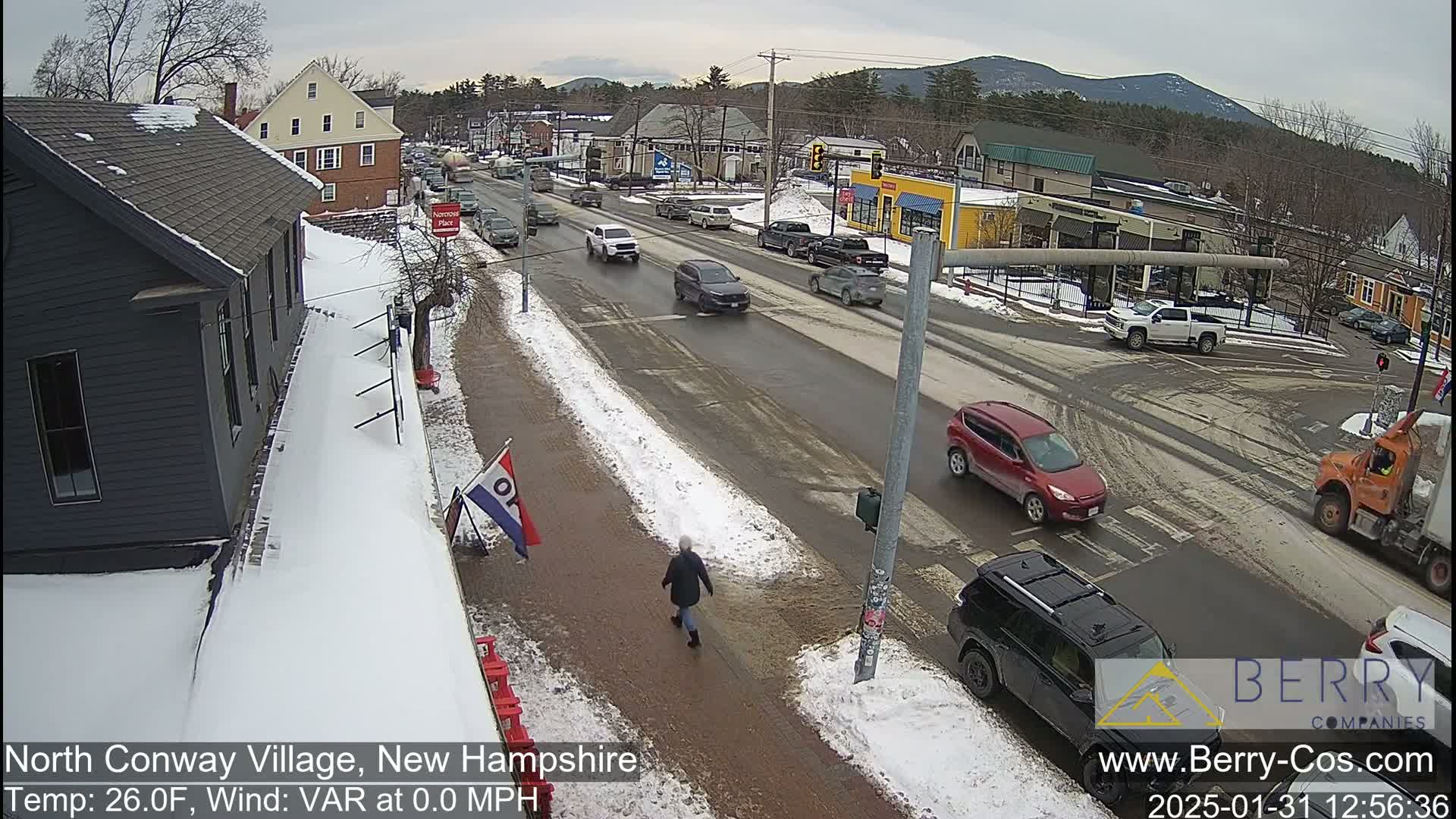 A snow-covered street scene shows several vehicles at an intersection in front of various buildings, with a person walking on a snow-covered sidewalk, under an overcast sky.